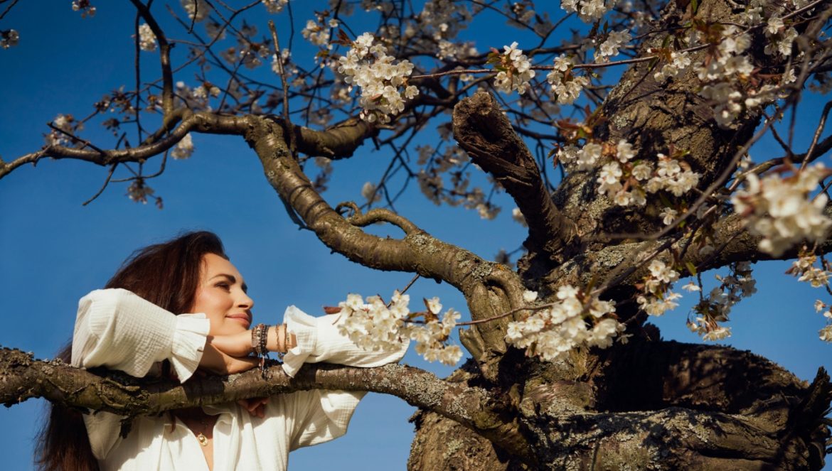Afirmacje Agnieszki Maciąg - pozytywne myśli i słowa to piękne życie. Agnieszka Maciąg opiera się rękami o gałąź drzewa z wiosennymi kwiatami.