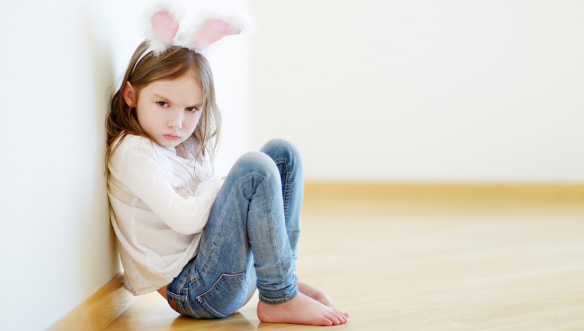 Jak radzić sobie z agresją u dzieci? Zła i agresywna dziewczynka z pluszowymi uszami królika na głowie siedzi pod ścianą i patrzy w obiektyw złym wzrokiem.