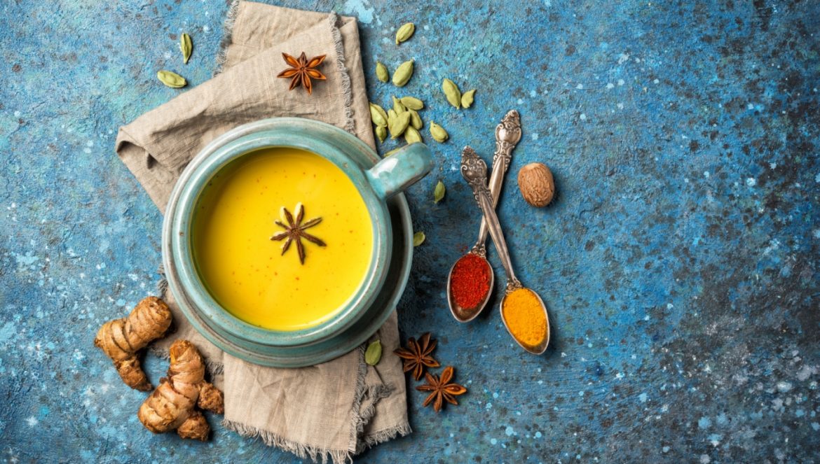 Jak zbudować odporność według ajurwedy? Miseczka napoju z kurkumy i goździków (złotego mleka) na niebieskim blacie, wokół leżą przyprawy, będące składnikami napoju.