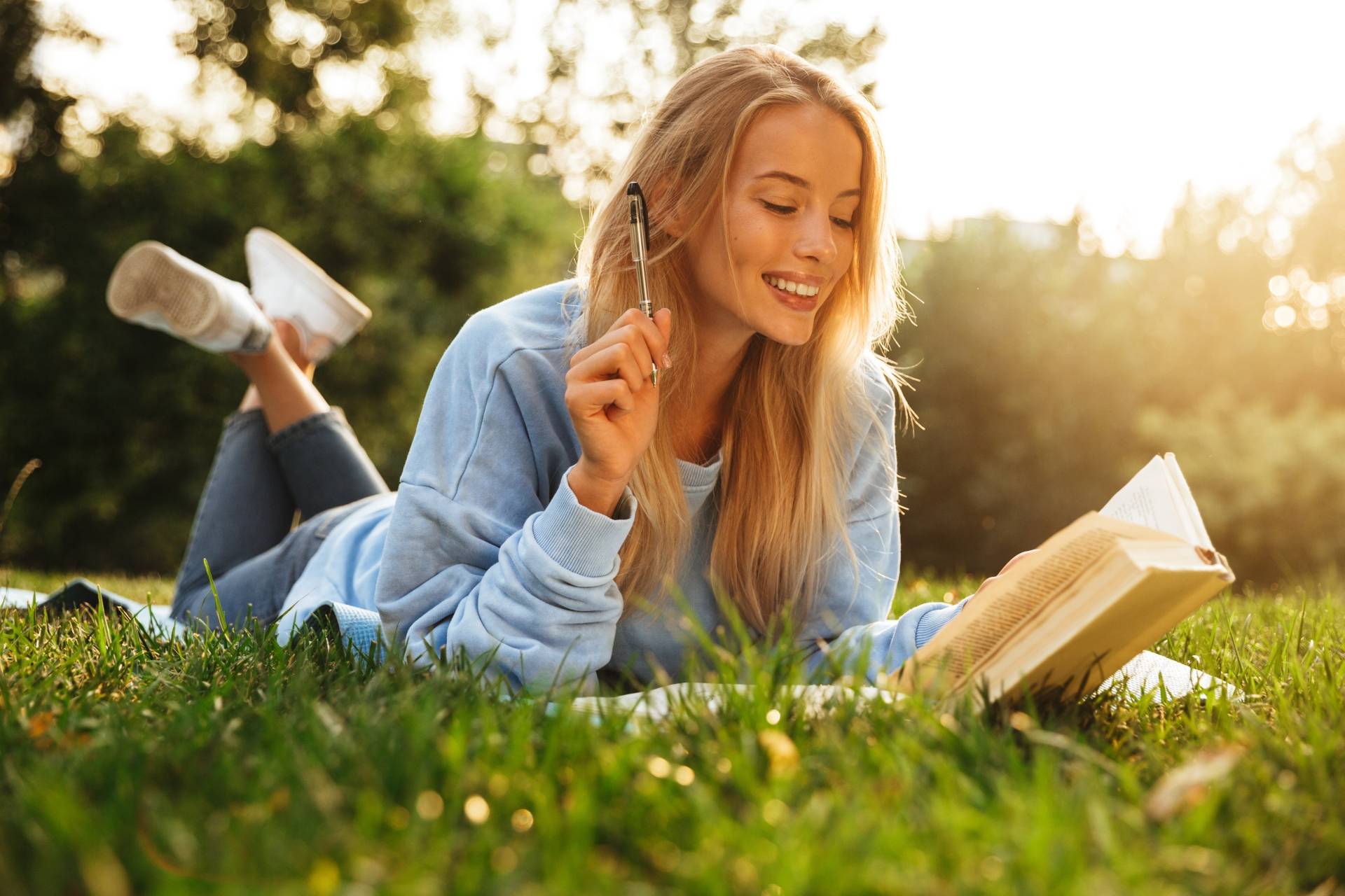 Alergia na pyłki traw i drzew. Młoda uśmiechnięta kobieta w niebieskiej bluzie leży na kocu i czyta książkę.
