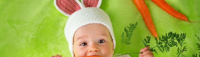 Alergia pokarmowa u dzieci - jak leczyć alergię jawną, a jak utajoną? Niemowlę w czapce z króliczymi uszami leży na zielonym kocyku i chrupie marchewkę.