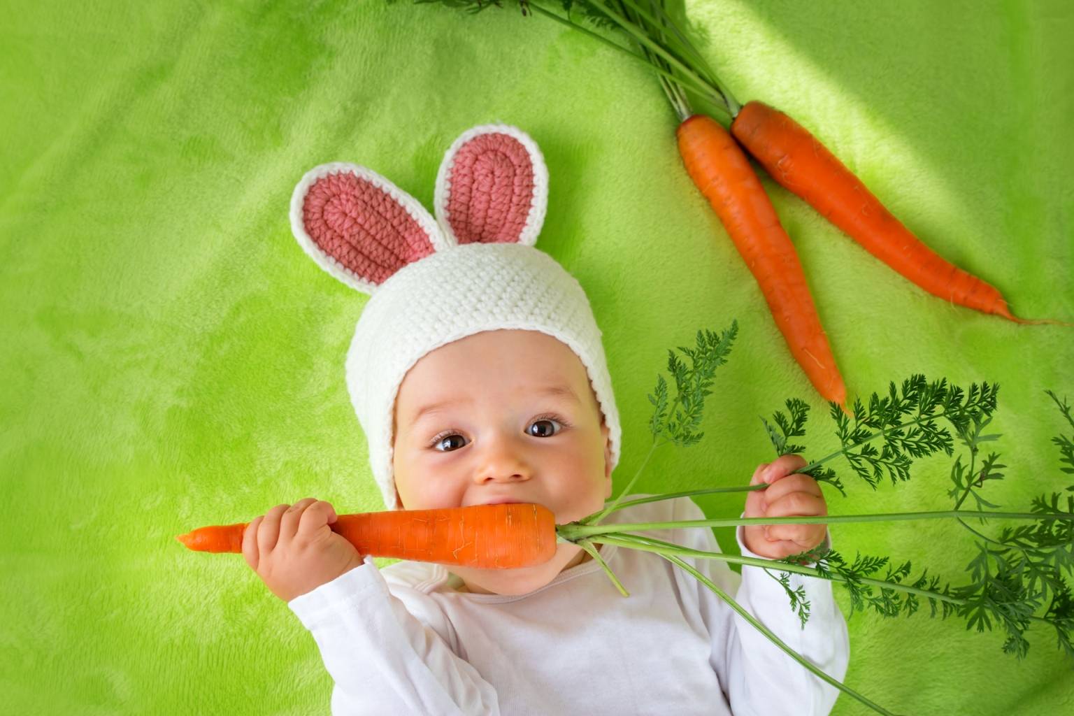 Alergia pokarmowa u dzieci - jak leczyć alergię jawną, a jak utajoną? Niemowlę w czapce z króliczymi uszami leży na zielonym kocyku i chrupie marchewkę.