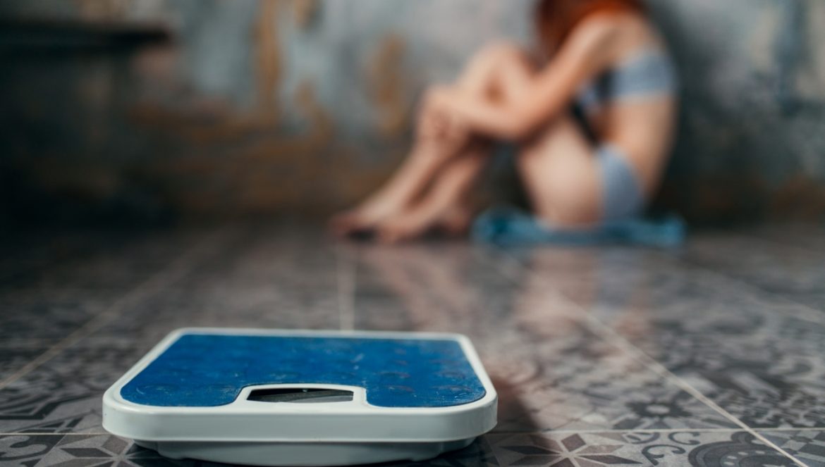 Anoreksja - jakie są jej przyczyny, objawy i jak ją leczyć? Anorektyczka siedzi w kącie, na pierwszym planie widać wagę.