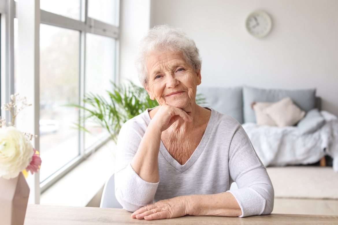 Kobieta seniorka siedzi przy stole w domu i uśmiecha się do obiektywu. Leki homeopatyczne dla seniorów.