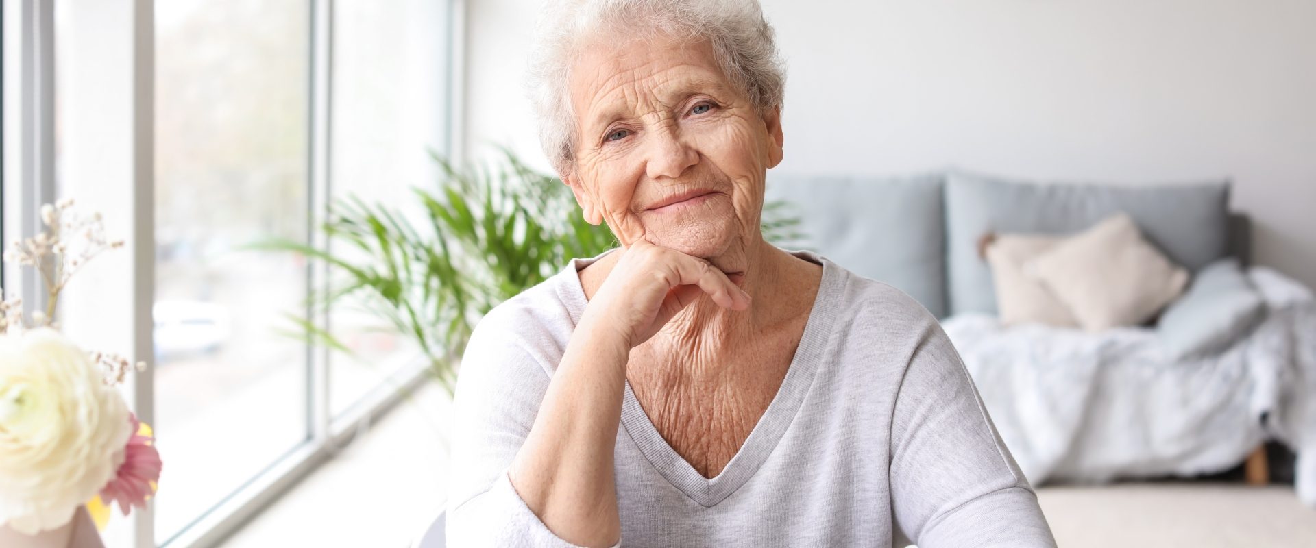 Kobieta seniorka siedzi przy stole w domu i uśmiecha się do obiektywu. Leki homeopatyczne dla seniorów.