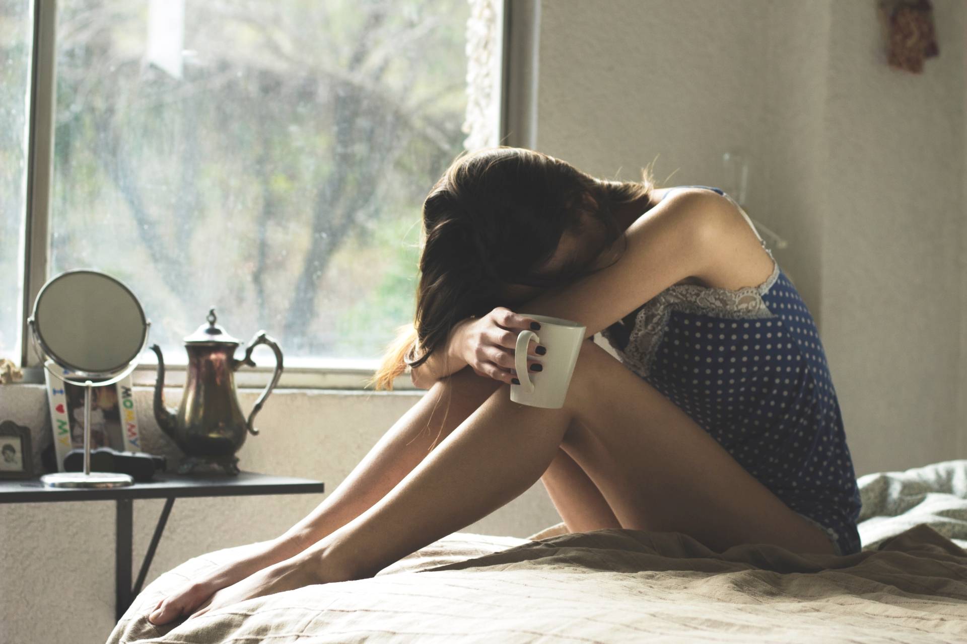 Jak poradzić sobie z traumą? Kobieta w depresji siedzi na łóżku w sypialni i opiera głowę o kolana, w ręku trzyma kubek.