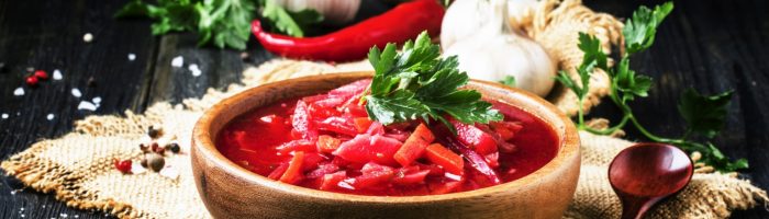 Przepis na zakwas buraczany - jak zrobić zakwas z buraków? Przepis i właściwości. Barszcz w drewnianej misce na lnianej ściereczce, w tle leży czosnek, natka i papryczka.