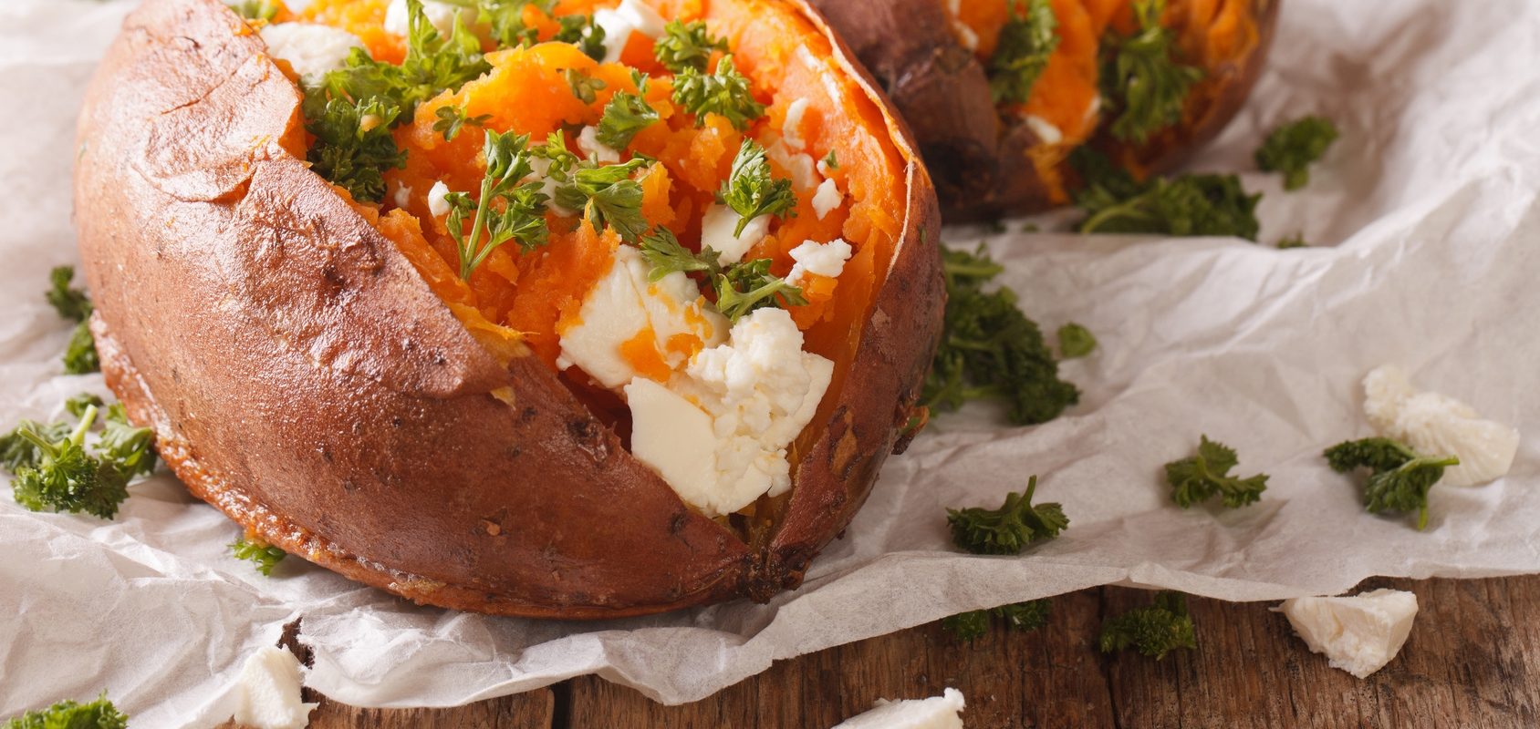 Bataty - dlaczego warto jej jeść? Przepisy z batatów. Słodkie ziemniaki z fetą i natką na białym papierze do pieczenia i drewnianym blacie.