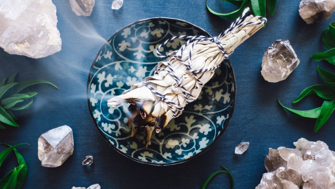 Biała szałwia do okadzania domu dymi na talerzu w niebieskie etniczne wzorki na granatowym blacie. Wokół leżą zielone listki ziół.