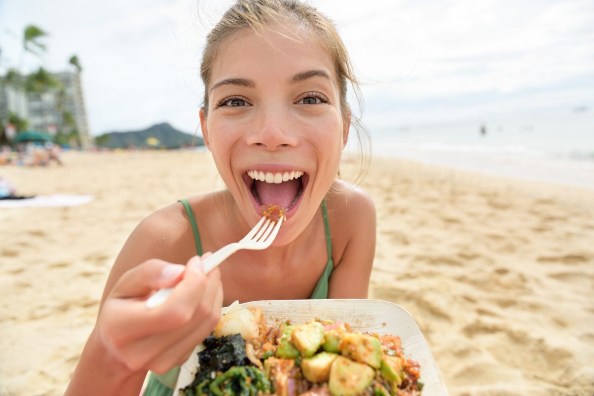Biegunka podróżnych - jak się przed nią ustrzec i jak ją leczyć? Uśmiechnięta dziewczyna w zielonej sukience na plaży je regionalną potrawę wziętą na wynos z lokalnego baru.