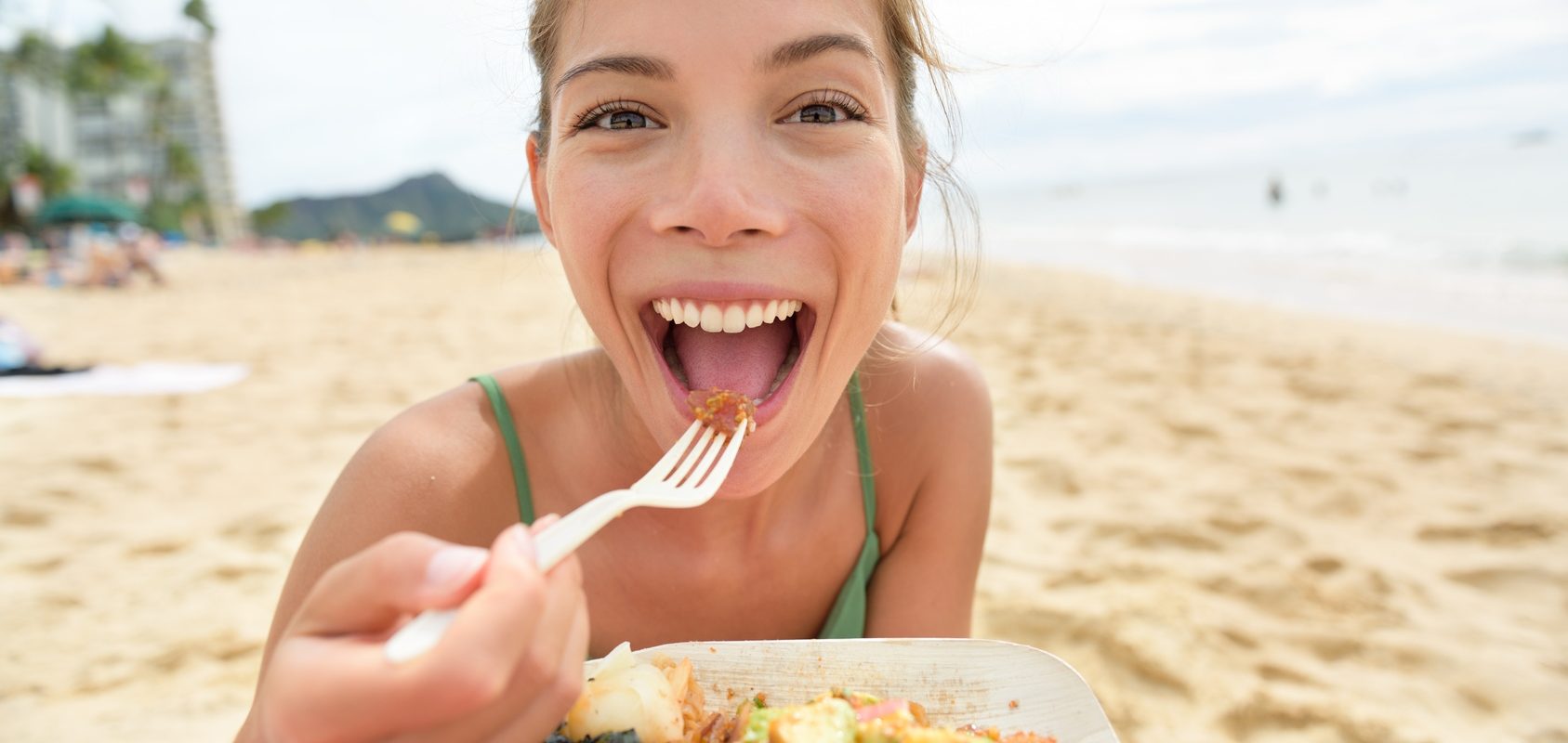 Biegunka podróżnych - jak się przed nią ustrzec i jak ją leczyć? Uśmiechnięta dziewczyna w zielonej sukience na plaży je regionalną potrawę wziętą na wynos z lokalnego baru.