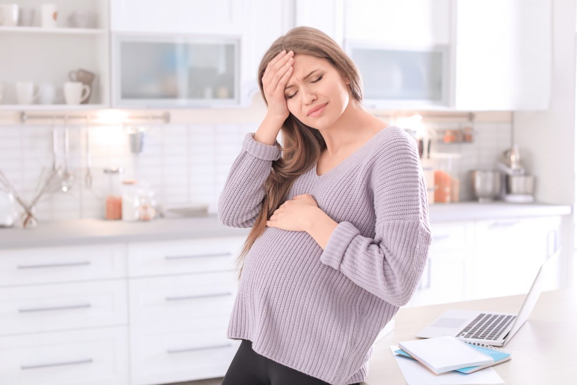 Ból głowy w ciąży - jak sobie z nim poradzić? Kobieta w zaawansowanej ciąży trzyma się za bolącą głowę, stojąc w kuchni.