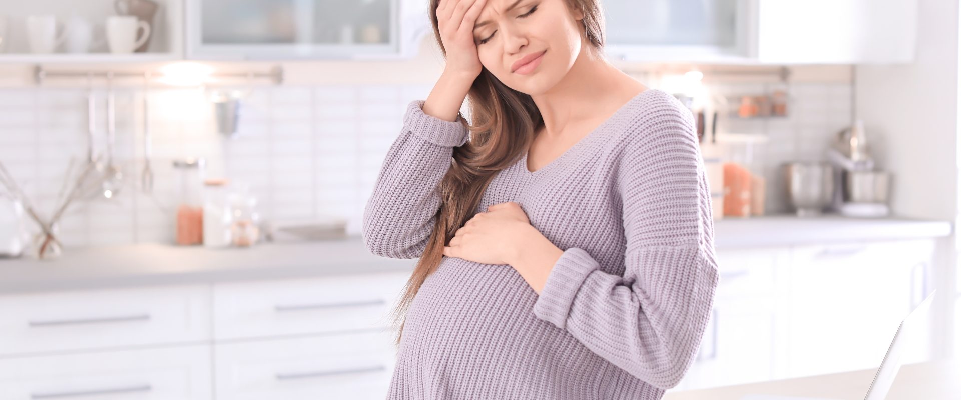 Ból głowy w ciąży - jak sobie z nim poradzić? Kobieta w zaawansowanej ciąży trzyma się za bolącą głowę, stojąc w kuchni.