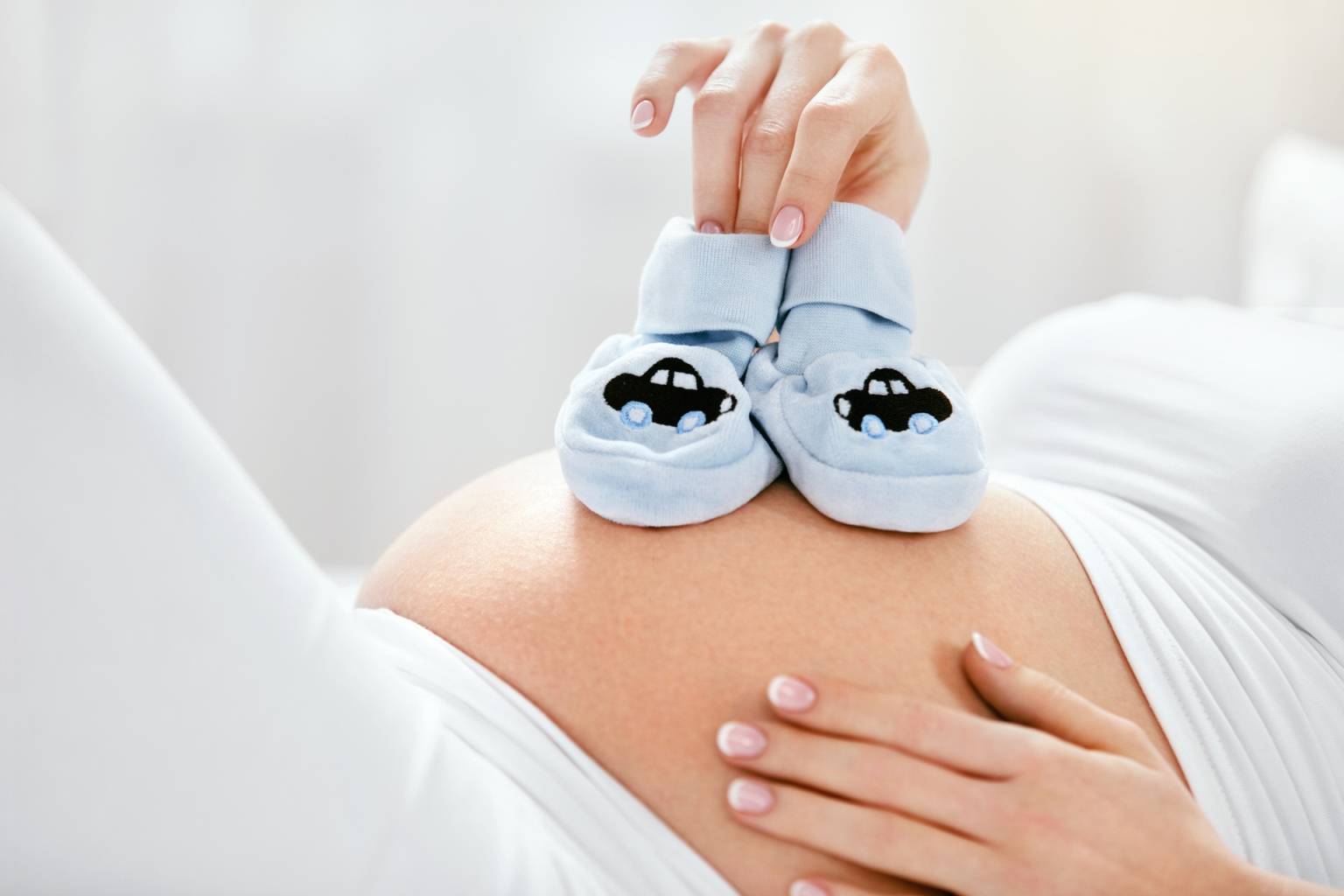 Jak przygotować się na narodziny dziecka? Homeopatia przed porodem i na połóg. Kobieta kładzie małe buciki niemowlęce na brzuch w zaawansowanej ciąży.