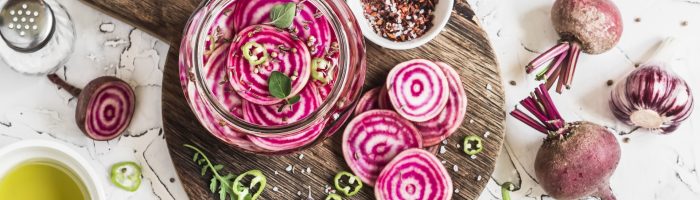 Najlepsze przepisy na kiszonki: kapusta kiszona, kiszone buraki, kiszone jabłka, kiszone cytryny, kimchi. Pokrojone w talarki buraki do zakiszenia na drewnianej desce na białym blacie, wokół przyprawy i zioła.