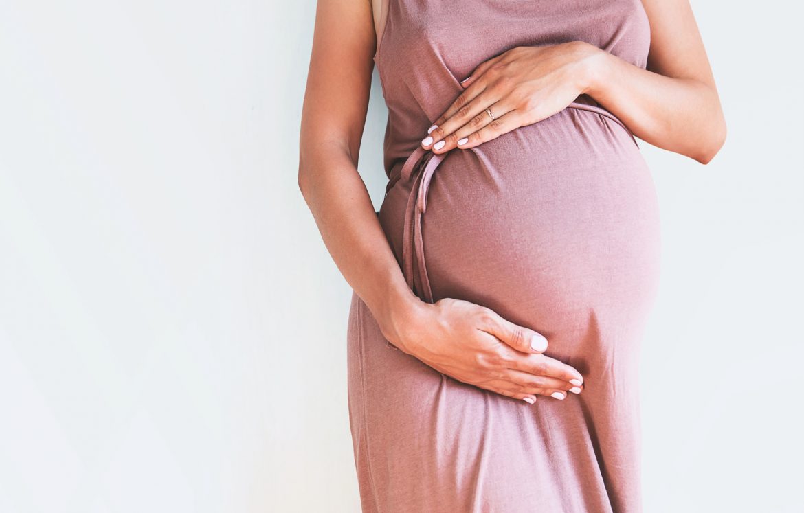 Cesarskie cięcie czy poród naturalny? Jakie są zalety porodu siłami natury i jak zadbać o noworodki urodzone przez CC? Kobieta w zaawansowanej ciąży w różowej sukience dotyka brzucha.