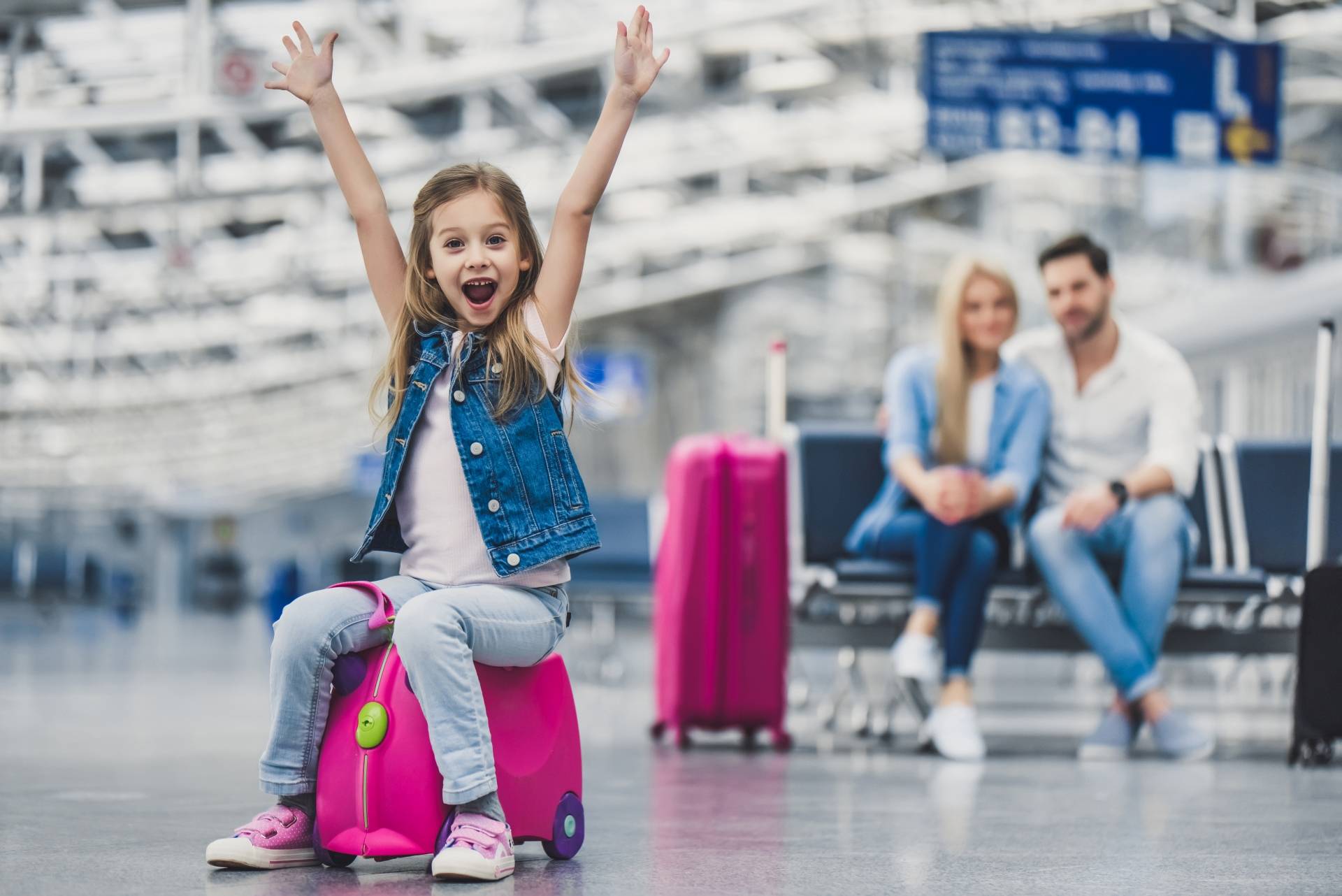 Sposoby na chorobę lokomocyjną. Dziewczynka siedzi na swojej różowej walizeczce na lotnisku, w tle na ławce siedzą objęci rodzice.