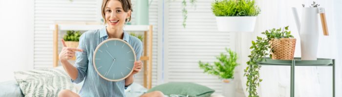 Chronobiologia - na czym polega życie we własnym rytmie? Czym są chronotypy i rytm dobowy? Kobieta siedzi na łóżku w sypialni, otoczona roślinami i trzyma w ręku zegar ścienny.