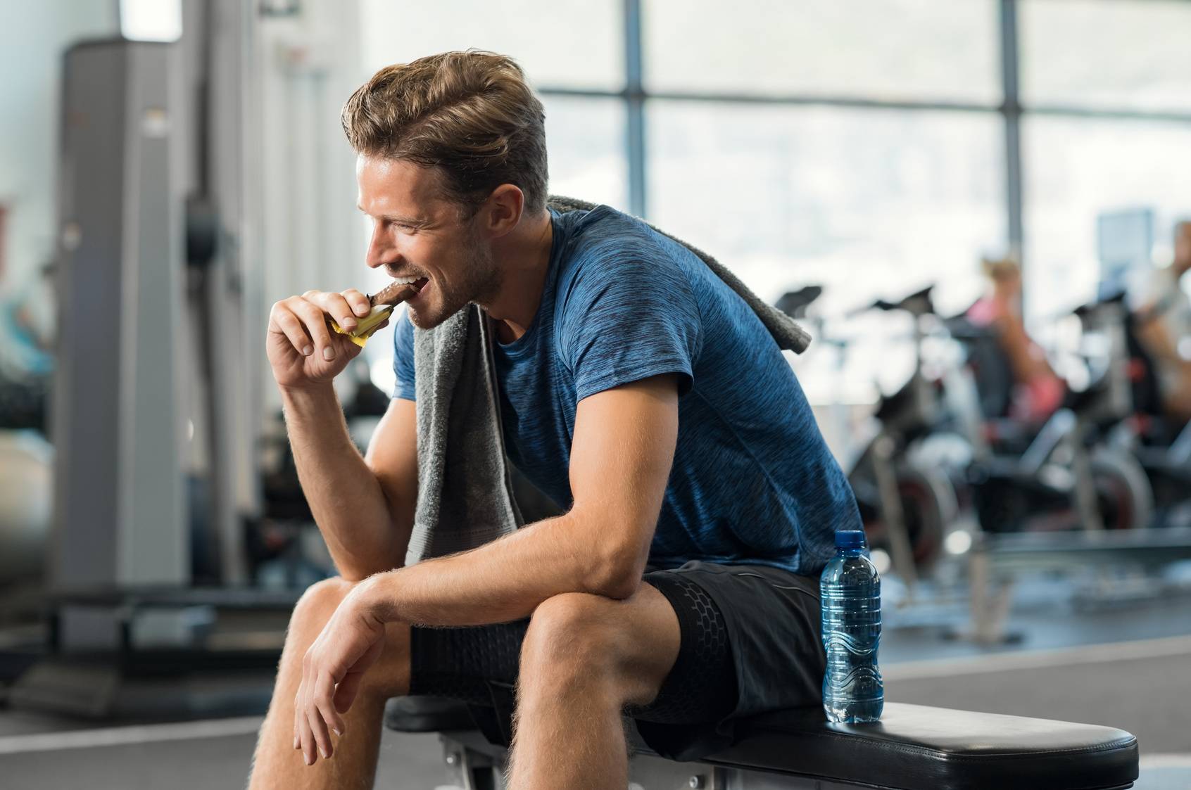 Posiłek potreningowy i posiłek przed treningiem - co i kiedy jeść? co jeść po treningu? Mężczyzna po treningu na siłowni je batonika proteinowego.
