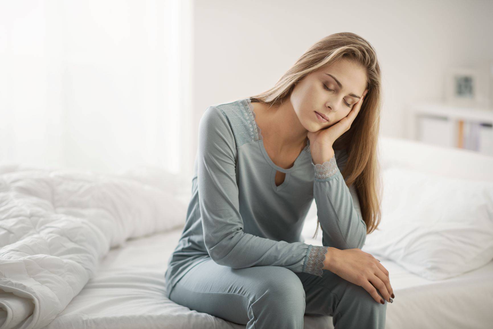 Homeopatia na infekcję. Kto potrzebuje leku Ferrum phosphoricum i na co on pomaga? Zmęczona chora kobieta siedzi w piżamie na łóżku w sypialni i podpiera ręką głowę.