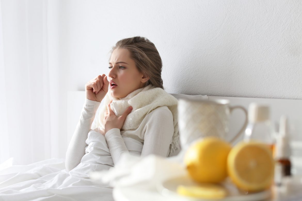 Co na kaszel? Sposoby na kaszel suchy i mokry. Jakie leki homeopatyczne zastosować przy kaszlu? Młoda kobieta opatulona swetrem i szalikiem siedzi w łóżku pod kołdrą i kaszle.