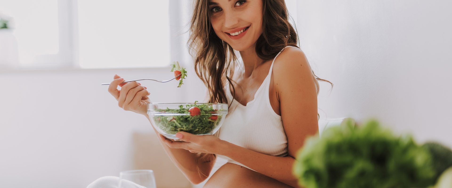 Cukrzyca ciążowa - jak leczyć cukrzycę w ciąży? Młoda kobieta w ciąży je sałatkę i uśmiecha się do aparatu.