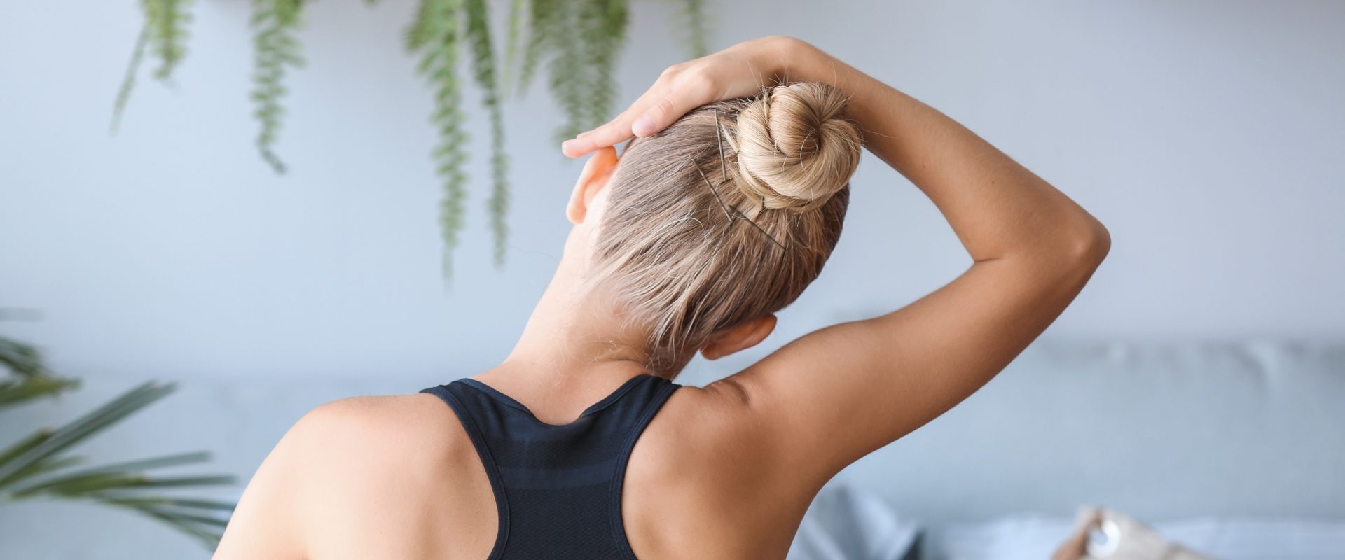 Ćwiczenia na kręgosłup szyjny. Kobieta w czarnym topie z włosami związanymi w kok siedzi zwrócona tyłem do aparatu i rozciąga szyję.