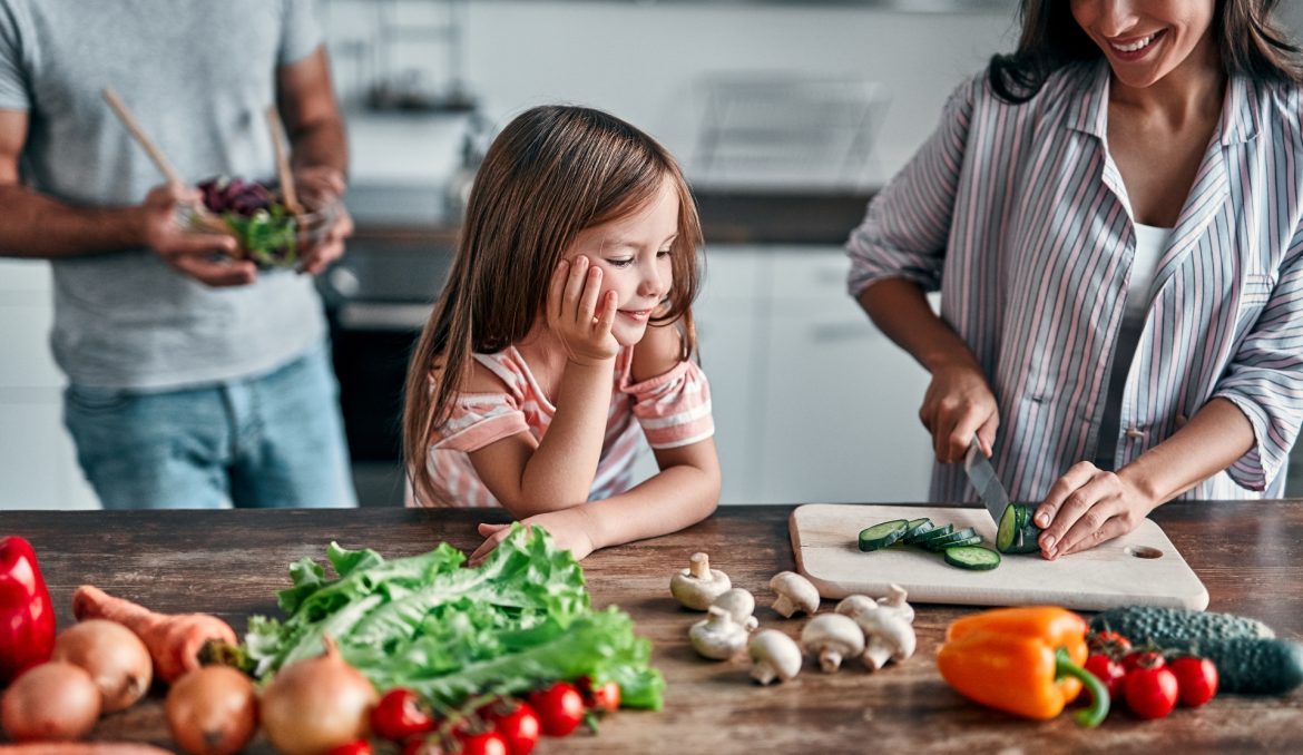 Cynk i selen na odporność - gdzie szukać w diecie tych cennych składników i czy faktycznie wzmacniają układ odpornościowy? Dziewczynka przygląda się jak mama kroi warzywa w kuchni, w tle stoi ojciec.