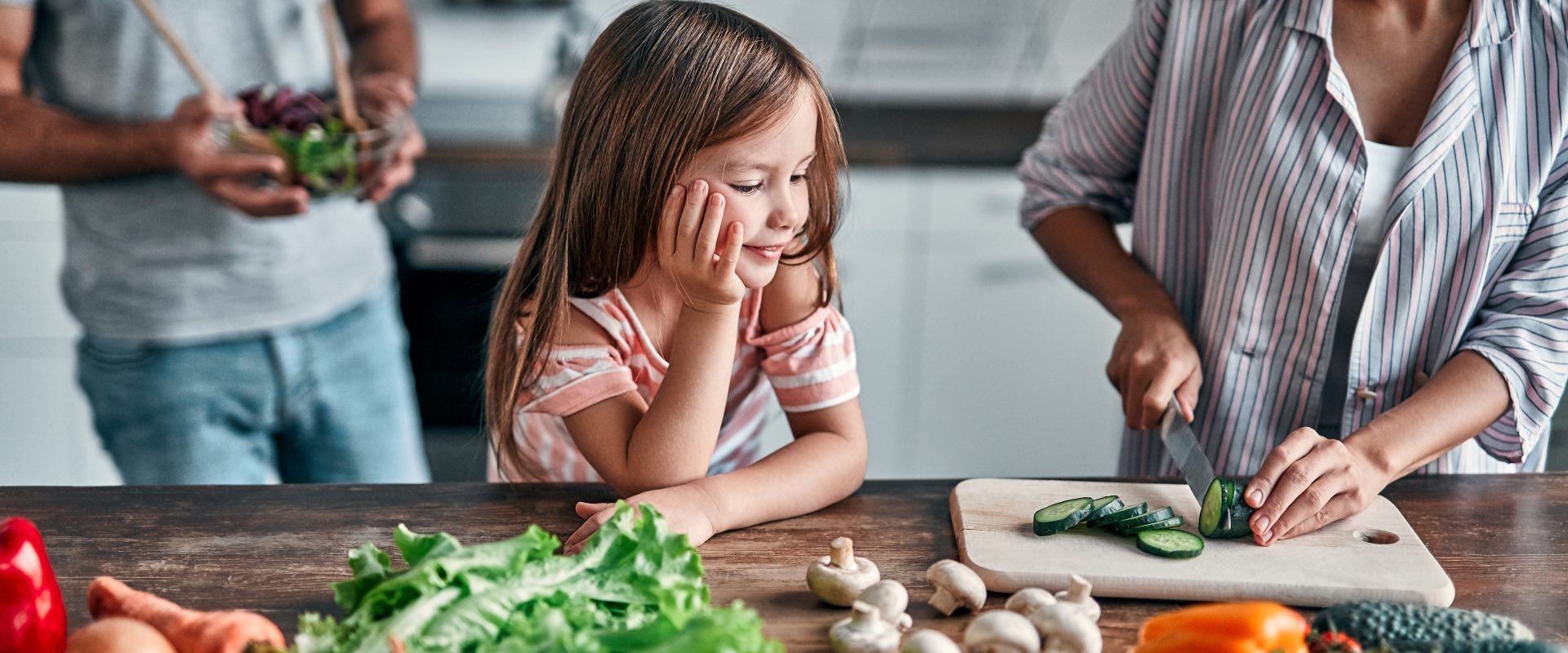 Cynk i selen na odporność - gdzie szukać w diecie tych cennych składników i czy faktycznie wzmacniają układ odpornościowy? Dziewczynka przygląda się jak mama kroi warzywa w kuchni, w tle stoi ojciec.