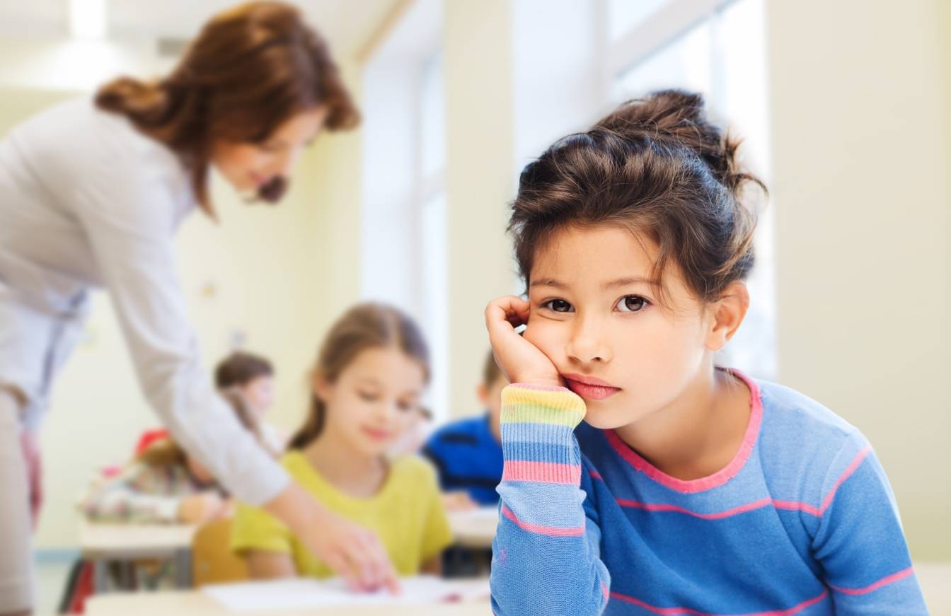 Depresja u dzieci - jakie są objawy depresji u dzieci, przyczyny i jakie wdrożyć leczenie? Smutna dziewczynka opiera głowę na ręku, siedząc w ławce w klasie.