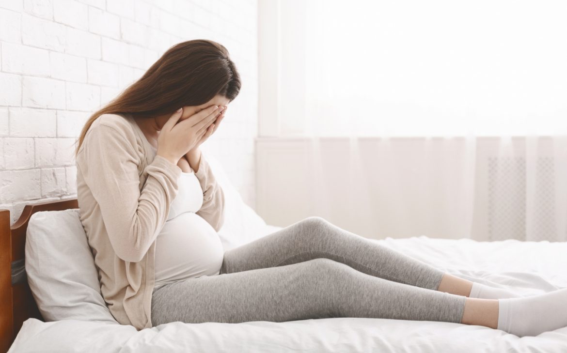 Depresja w ciąży - objawy, przyczyny, leczenie, skutki. Jak poradzić sobie z depresją w ciąży? Załamana kobieta ciężarna siedzi na łóżku w sypialni i ukrywa twarz w dłoniach.