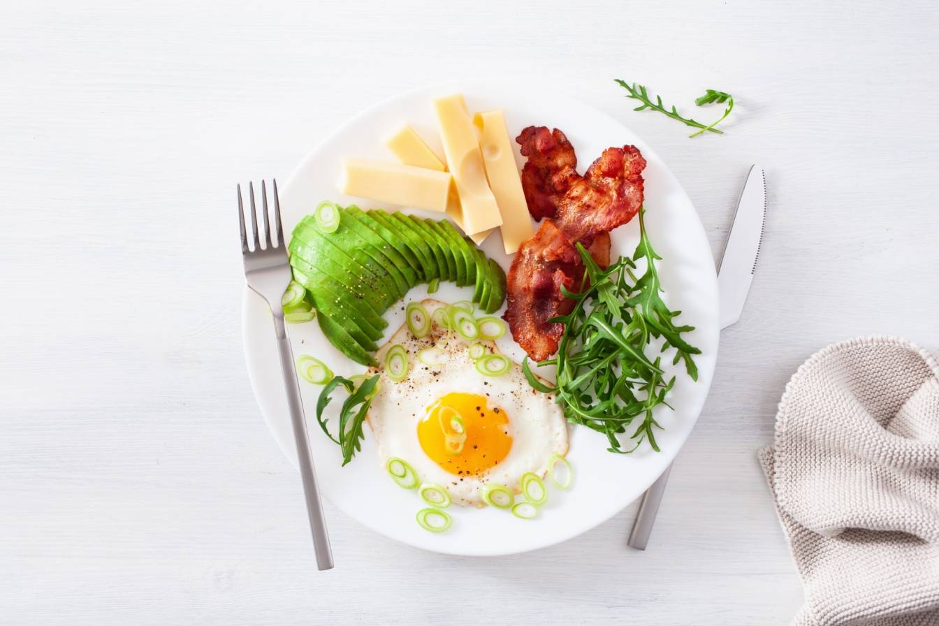 Co jeść na diecie ketogenicznej? Zdrowe śniadanie na diecie keto - jajka sadzone, bekon, awokado, ser żółty i rukola na białym talerzu.