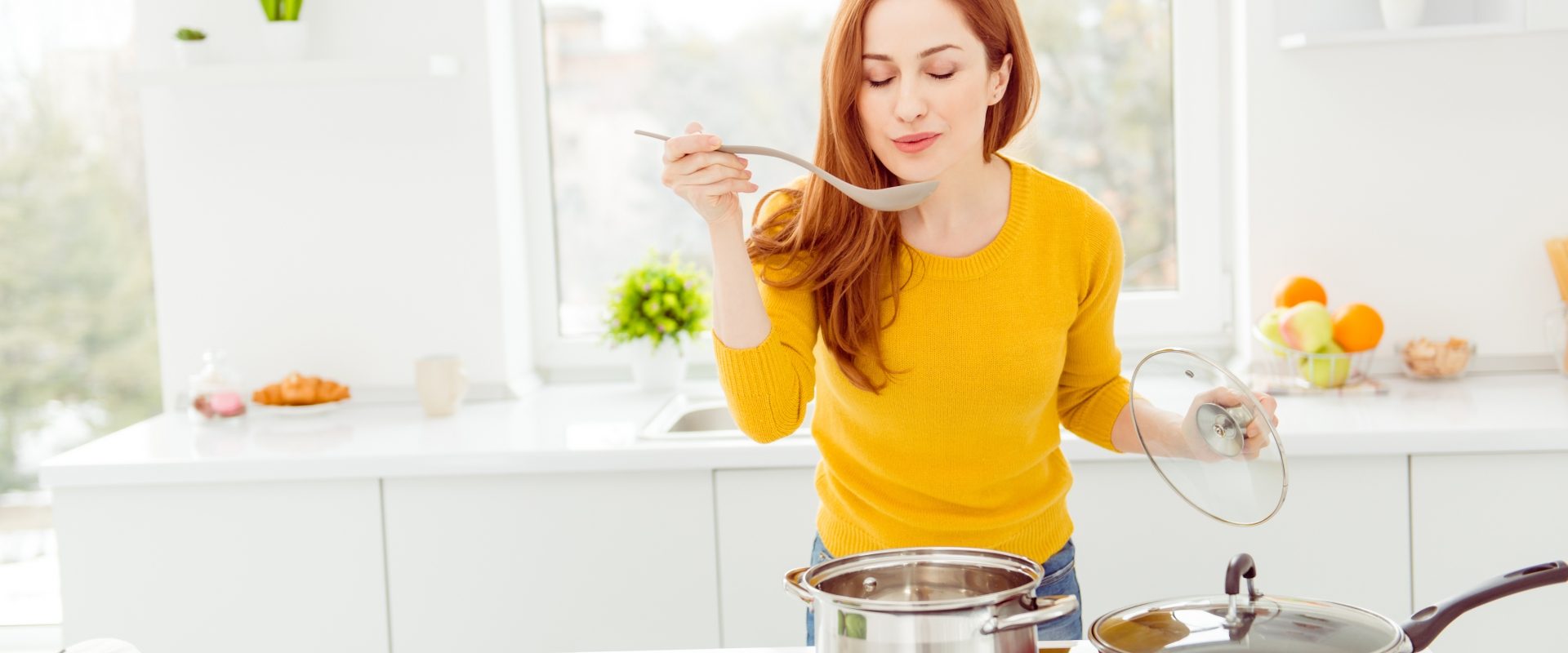 Dieta na alergię. Co powinni jeść alergicy? Rudowłosa kobieta w żółtym sweterku smakuje ugotowanej potrawy w swojej kuchni.