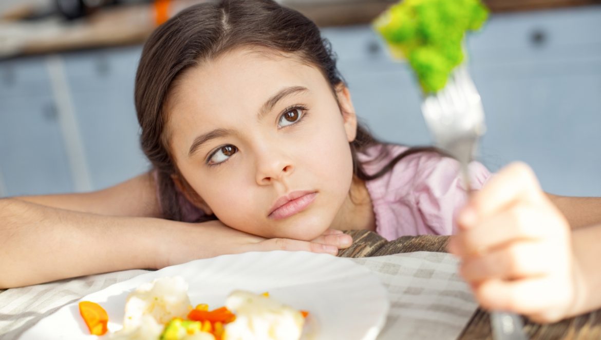 Autyzm i dieta, dieta w autyzmie - czy sposób żywienia może zwiększyć komfort życiowy dziecka opowiada dietetyczka kliniczna dr. n. med. Justyna Jessa (Mama dietetyk). Na zdjęciu autystyczna dziewczynka siedzi przy stole nad talerzem warzyw i przygląda się z oddali brokułowi nadzianemu na widelec.