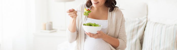 Dieta w ciąży - co najlepiej jeść? Kobieta w ciąży je sałatkę, siedząc na łóżku.