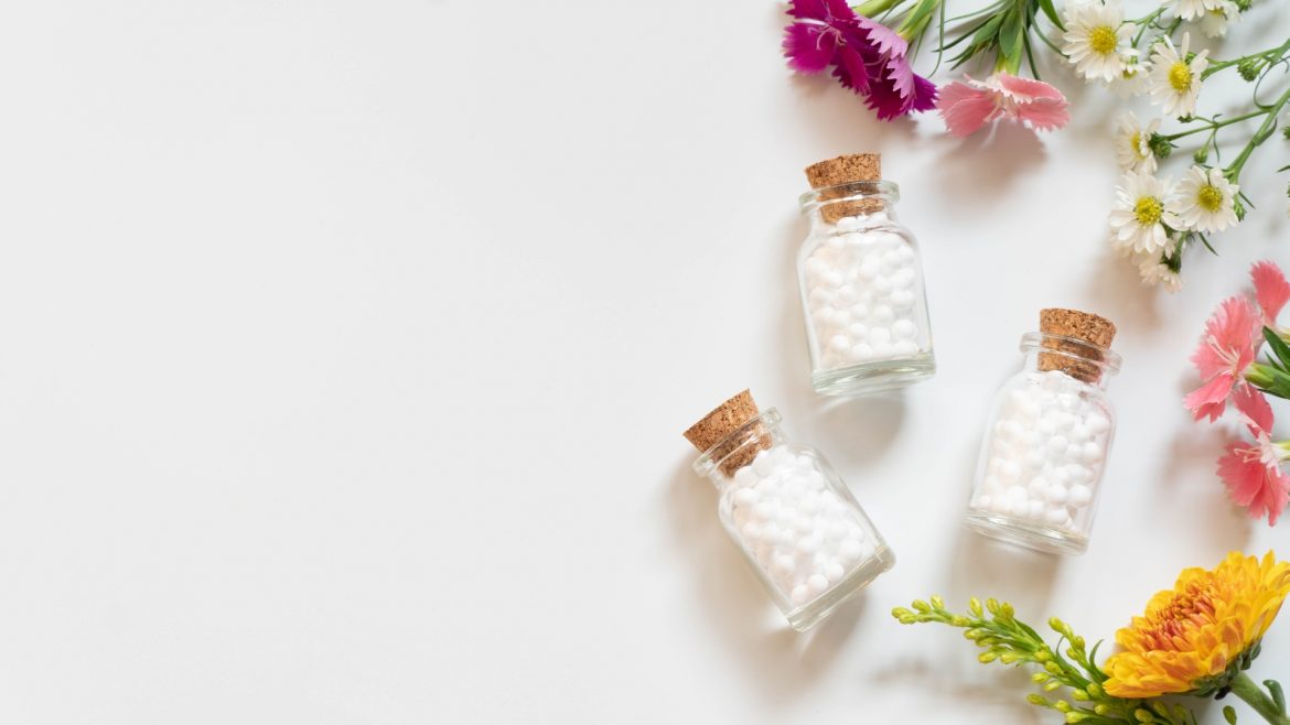 Homeopatia na niestrawność. Leki homeopatyczne w fiolkach, leżące na płasko na białym blacie. Obok leżą kwiaty.