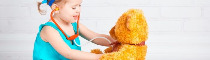 Rotawirus u dzieci - jak poradzić sobie z grypą żołądkową? Dziewczynka w niebieskim ubranku bawi się w doktora z pluszowym misiem - osłuchuje go stetoskopem.