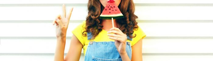 Jak nie chorować na wakacjach? Młoda dziewczyna ubrana w żółtą bluzkę, okulary przeciwsłoneczne i ogrodniczki je kawałek arbuza nabitego na patyk i pokazuje na palcach znak victorii.