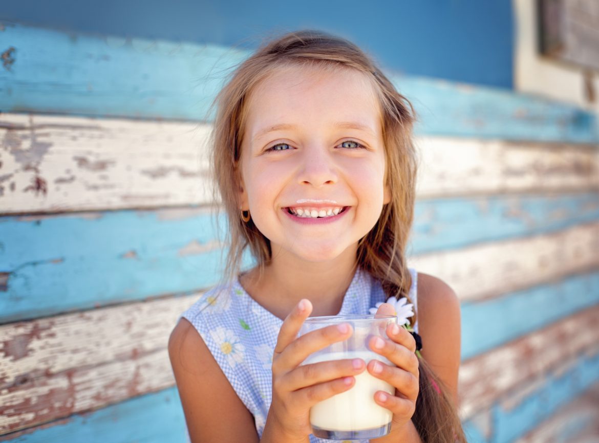 Mleko bez laktozy - pić czy nie pić? Co warto o nim wiedzieć? Uśmiechnięta dziewczynka z wąsami od mleka trzyma w dłoniach szklankę z mlekiem.