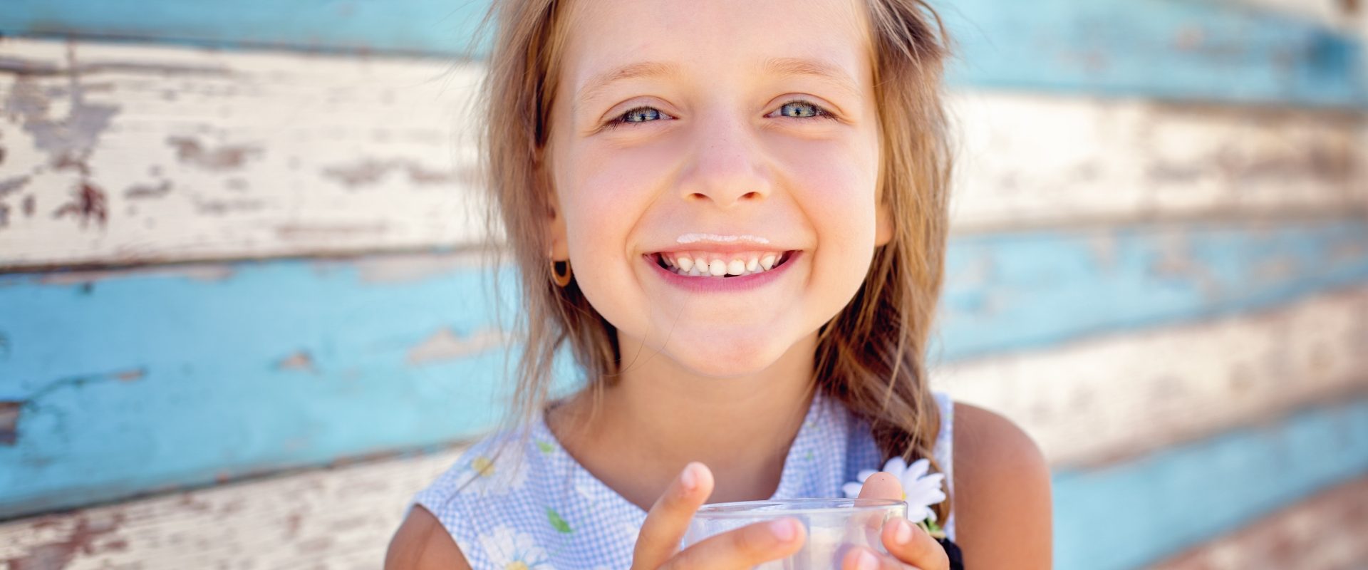 Mleko bez laktozy - pić czy nie pić? Co warto o nim wiedzieć? Uśmiechnięta dziewczynka z wąsami od mleka trzyma w dłoniach szklankę z mlekiem.