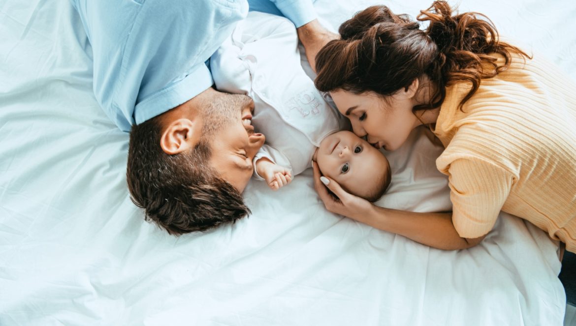 Ekorodzicielstwo - jak być ekorodzicem i nie zwariować? Rodzice tulą niemowlaka, leżąc na łóżku.