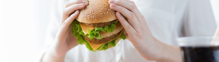 Jak poradzić sobie ze zgagą i refluksem? Co to jest refluks żołądkowo-przełykowy? Kobieta w białej bluzce je cheeseburgera z frytkami.