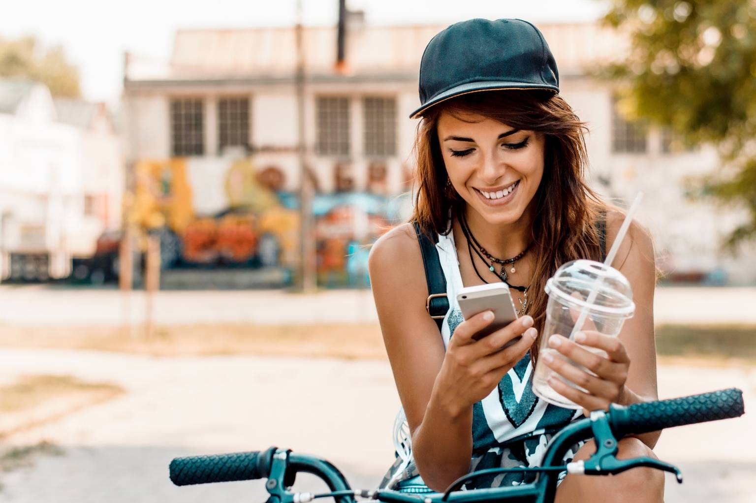 FOMO (Fear of missing out) - czym jest syndrom uzależnienia od informacji? Młoda dziewczyna w czapce z daszkiem siedzi na rowerze z telefonem i shakiem w rękach i czyta wiadomości.