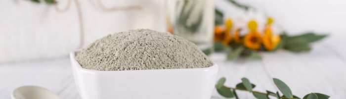 Glinka bentonitowa, sproszkowany bentonit w białej miseczce, wokół leżą gałązki roślin i kwiaty.