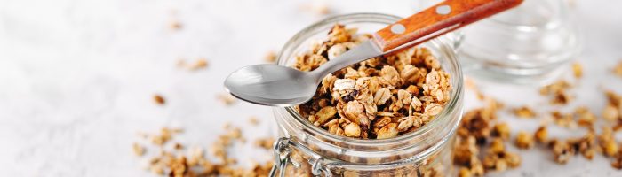 Domowa granola - przepis na granolę z płatkami owsianymi, orzechami, czekoladą. Granola wysypuje się ze słoika na biały kuchenny blat, na słoiku leży łyżeczka z drewnianą rączką.