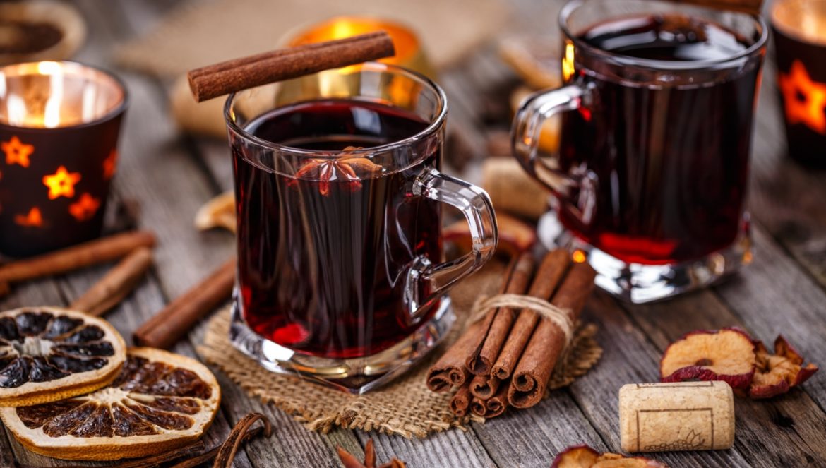Przepis na grzane wino - jak zrobić grzańca? Grzane wino w szklankach na stole obok korzennych przypraw, suszonych cytrusów i świątecznych akcentów.
