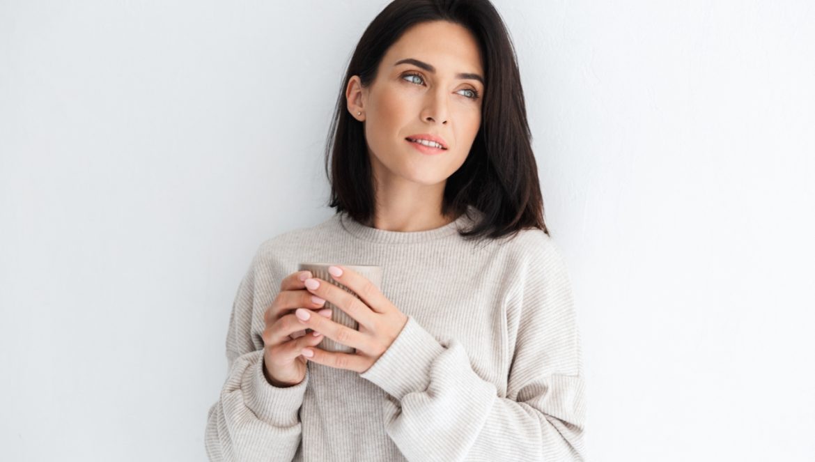 Hashimoto - objawy, przyczyny, diagnostyka i ciąża. Jak wykryć chorobę tarczycy? Atrakcyjna brunetka z kubkiem kawy w ręku stoi pod ścianą i spogląda w bok.