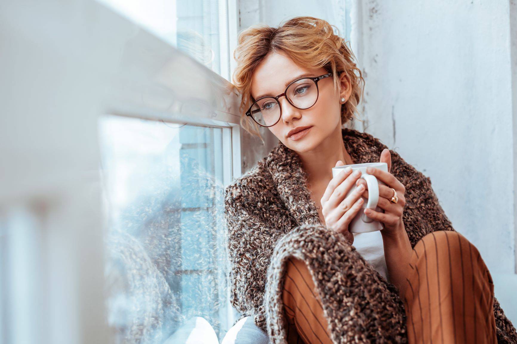 Jak żyć z chorobą Hashimoto? Smutna zamyślona kobieta siedzi z kubkiem herbaty na parapecie i wygląda przez okno.