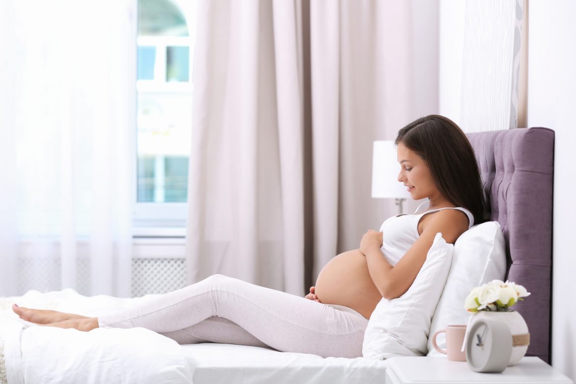 Hemoroidy w ciąży - jak sobie z nimi radzić? Kobieta w zaawansowanej ciąży siedzi na łóżku w sypialni i masuje się po brzuchu.