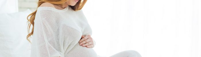 Ciąża po 40-tce - jak się dobrze przygotować do późnego macierzyństwa i jak pomóc sobie zajść w późną ciążę? Rudowłosa kobieta w zaawansowanej ciąży w białym luźnym ubraniu siedzi na łóżku na białej pościeli.